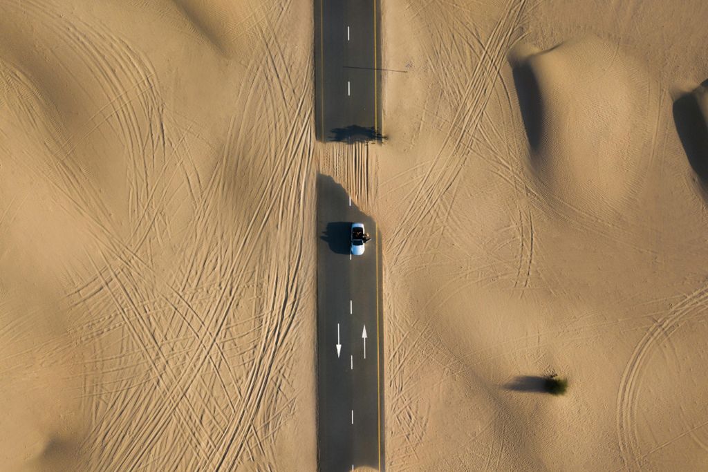 Aerial view of a car driving alone on a desert road with sand tracks on both sides.