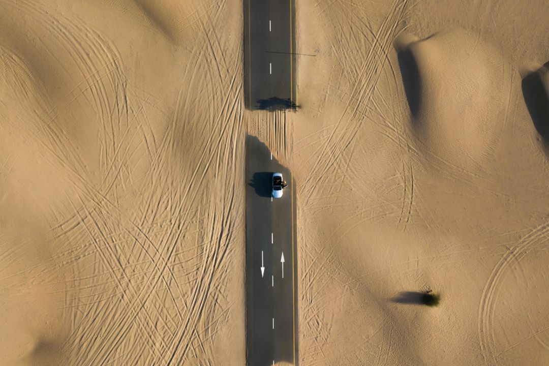 Aerial view of a car driving alone on a desert road with sand tracks on both sides.