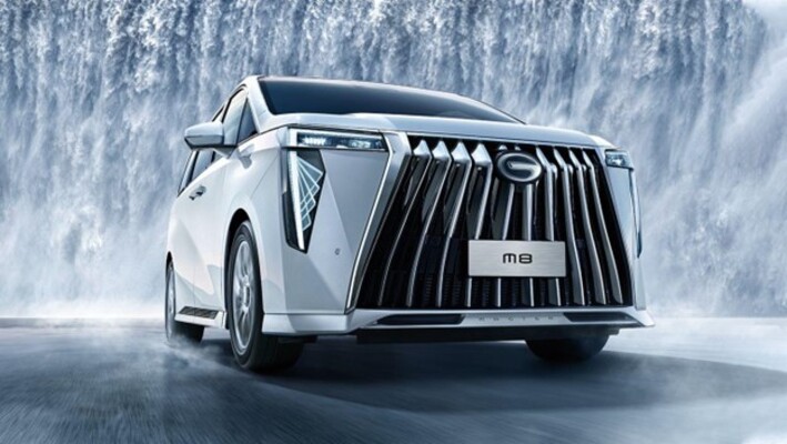 عرض أمامي لسيارة جي أيه سي M8 البيضاء تسير عبر منظر ضبابي مع شلال في الخلفية